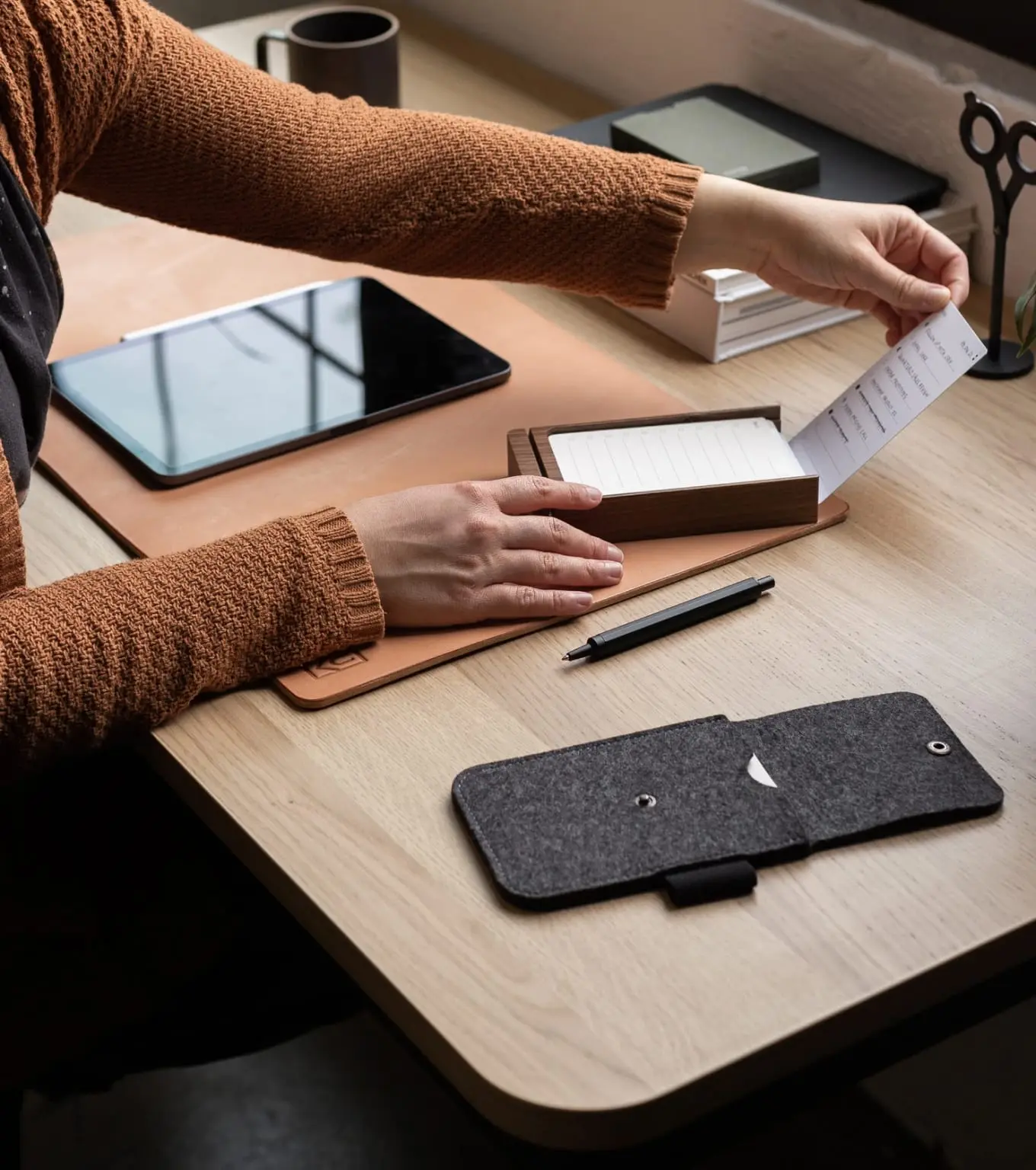 Visão aérea de uma pessoa organizada em sua mesa de trabalho, manuseando cartões de notas em um suporte de madeira, com um tablet e acessórios de escritório ao redor.